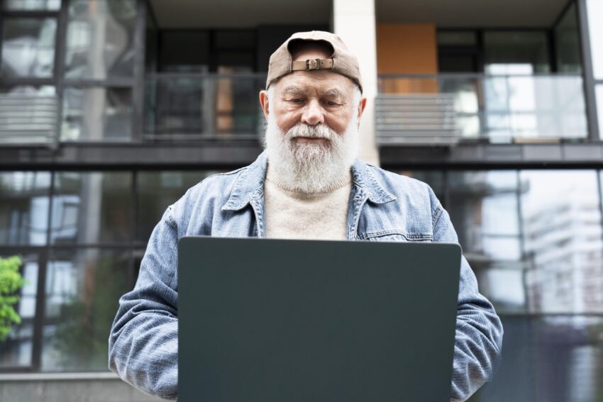 Homem de idade com chapéu para trás a utilizar um computador portátil na rua. Créditos: Freepik